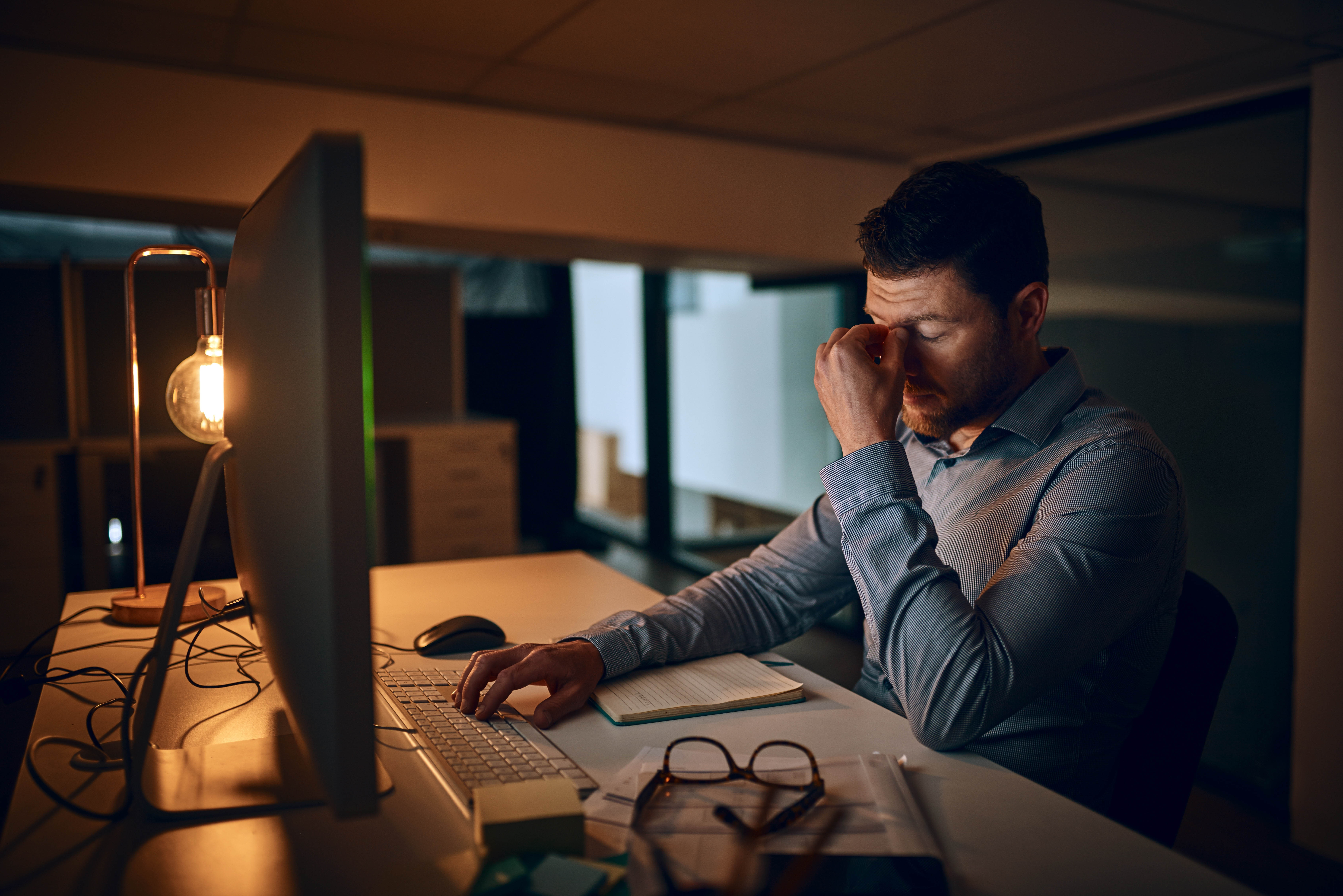 Man sitting in the dark in front of his computer, rubbing his eyes out of fatigue and tiredness